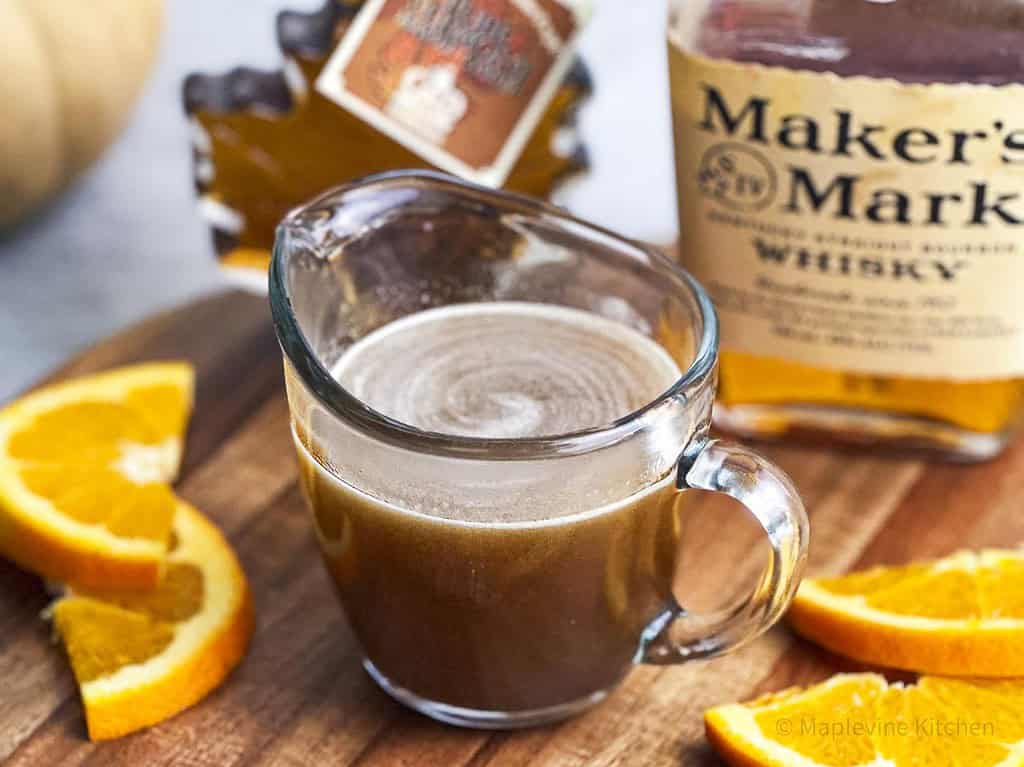 Maple bourbon glaze in a carafe on a wooden board with sliced oranges.