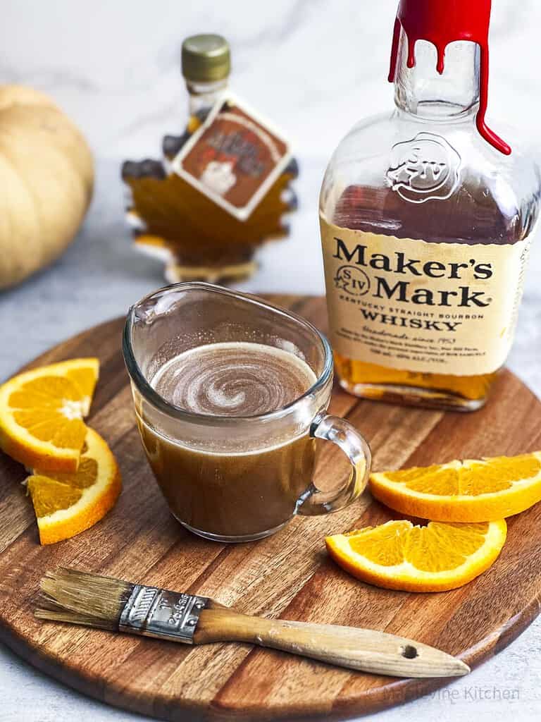 Maple bourbon glaze in a serving carafe with orange slices on the side.