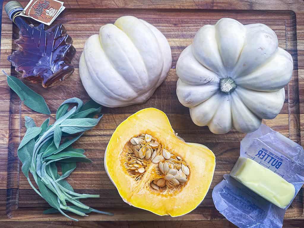 White acorn squash, sage leaves, maple syrup, and butter on a wooden cutting board.