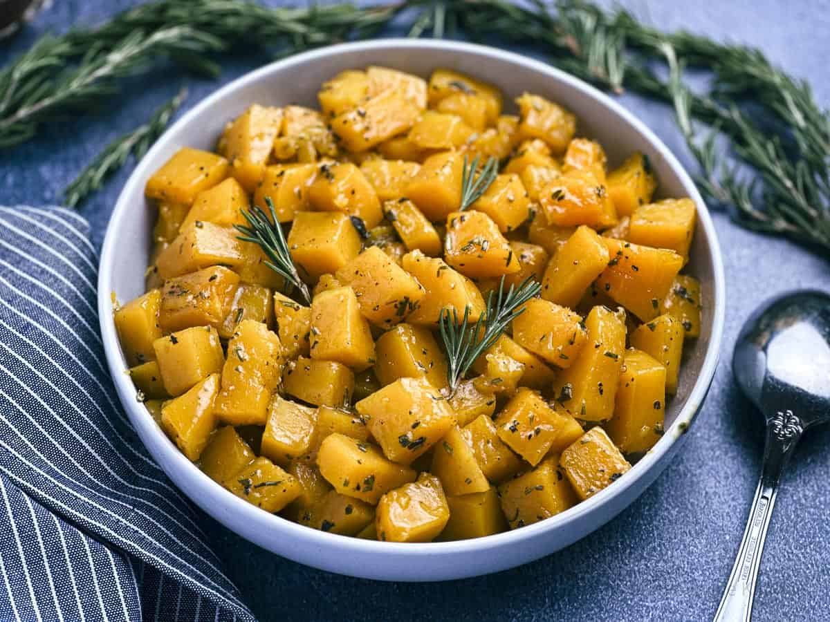 Apple cider glazed butternut squash in a bowl with a spoon and rosemary sprigs on the side.
