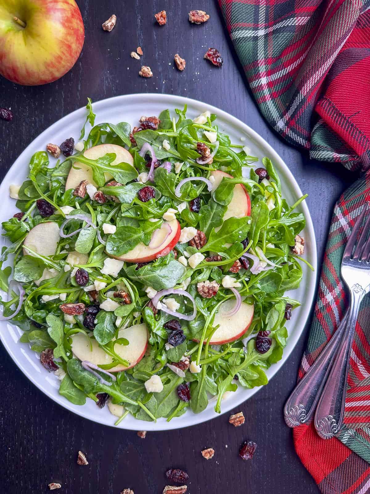 Arugula salad with apples, cheddar, pecans, and cranberries on a white plate.