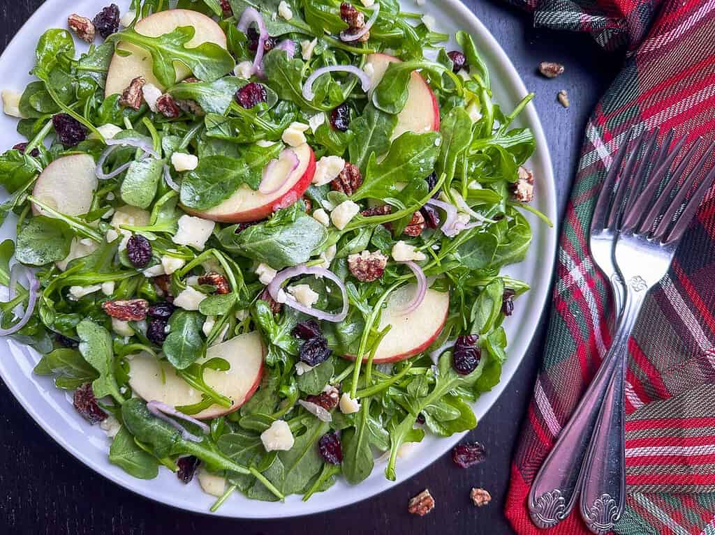 Arugula salad with apples, pecans, and cranberries on a white plate.