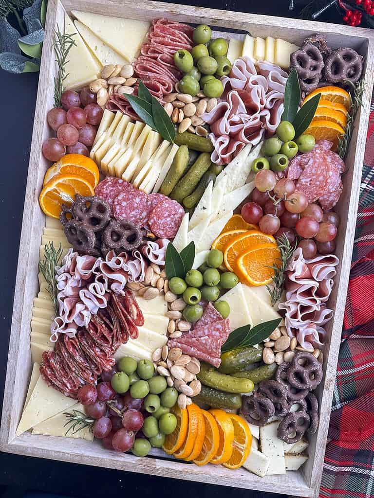 Holiday charcuterie board with salami, prosciutto, cheese, fruit, olives, pickles, and nuts arranged on a wooden tray.