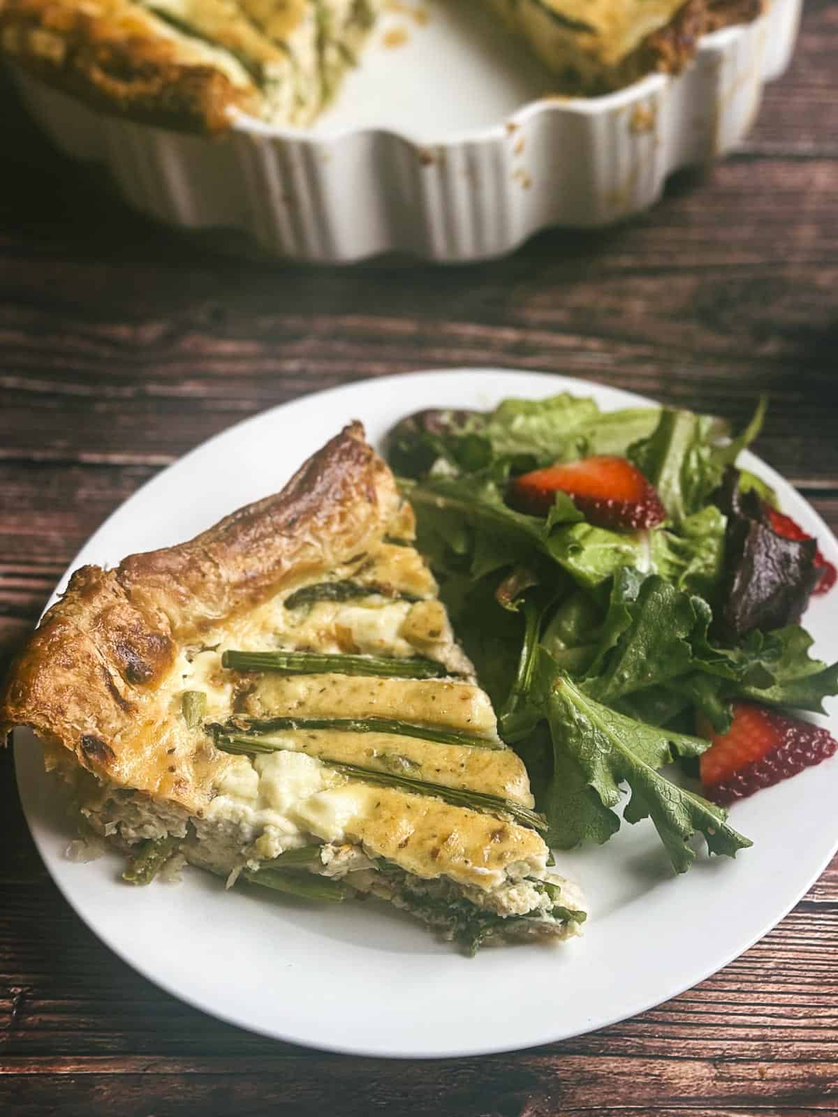 A slice of asparagus ricotta tart on white plate with strawberry spring mix salad.