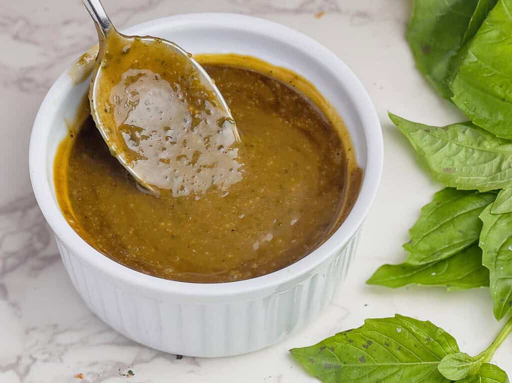 Balsamic vinaigrette in a ramekin with a spoon and basil leaves on the side.