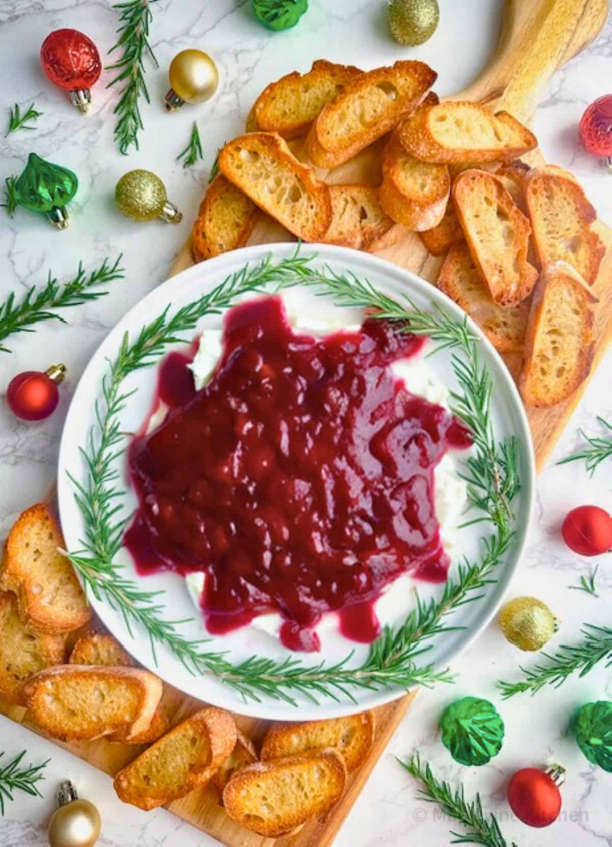 Cranberry whipped feta dip on a plate surrounded by crostini on a wood serving board with Christmas ornaments and rosemary.