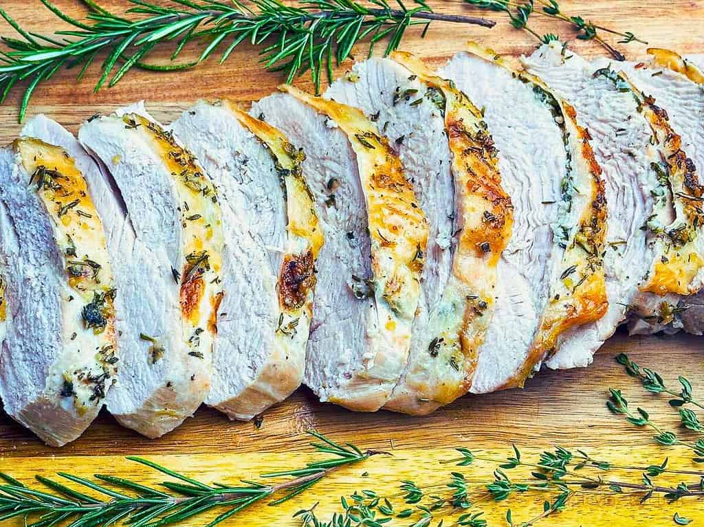 Roasted boneless turkey breast slices on a board surrounded by fresh herbs.