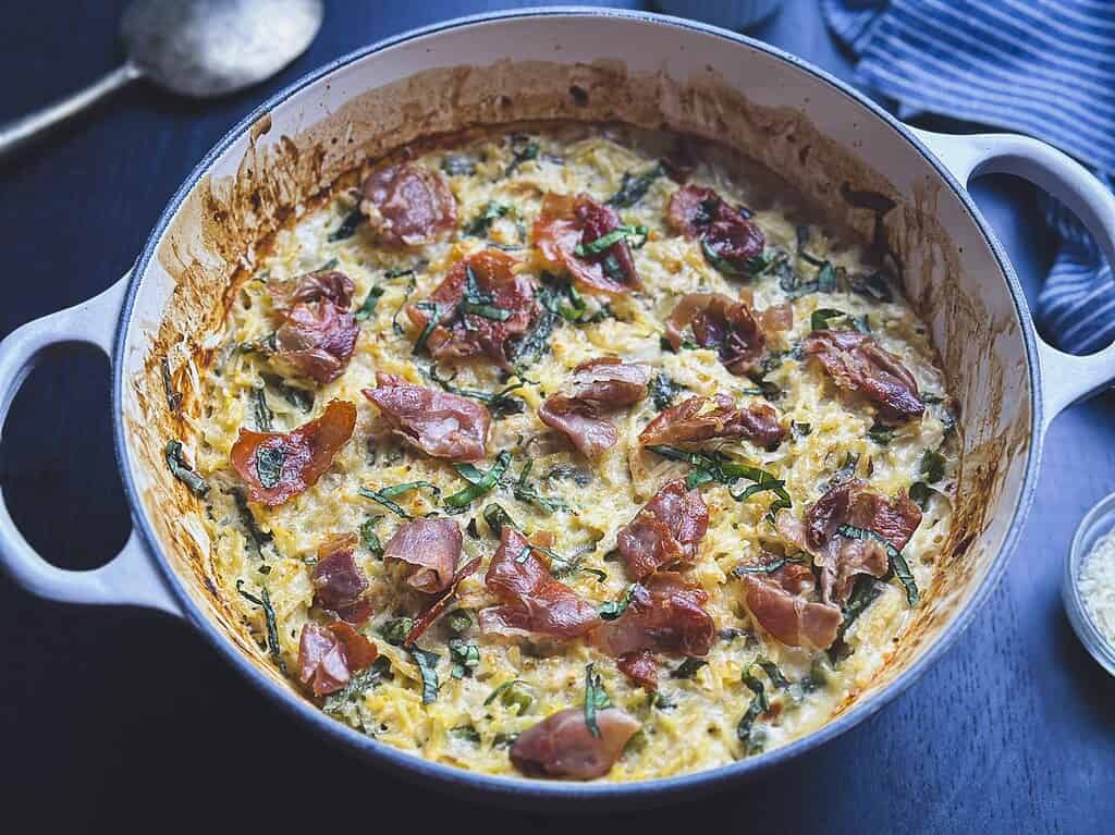 Creamy chicken orzo pasta bake in a Dutch oven with crispy prosciutto and basil on top.