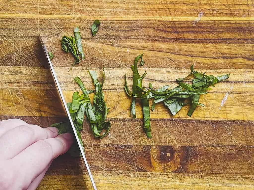 A hand and knife slicing basil on a cutting board using the chiffonade technique.