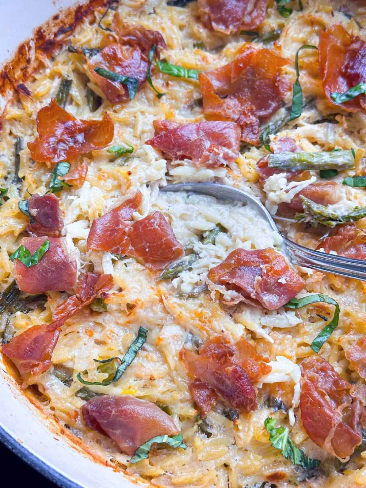 Close up of creamy chicken orzo with prosciutto on top with a spoon.