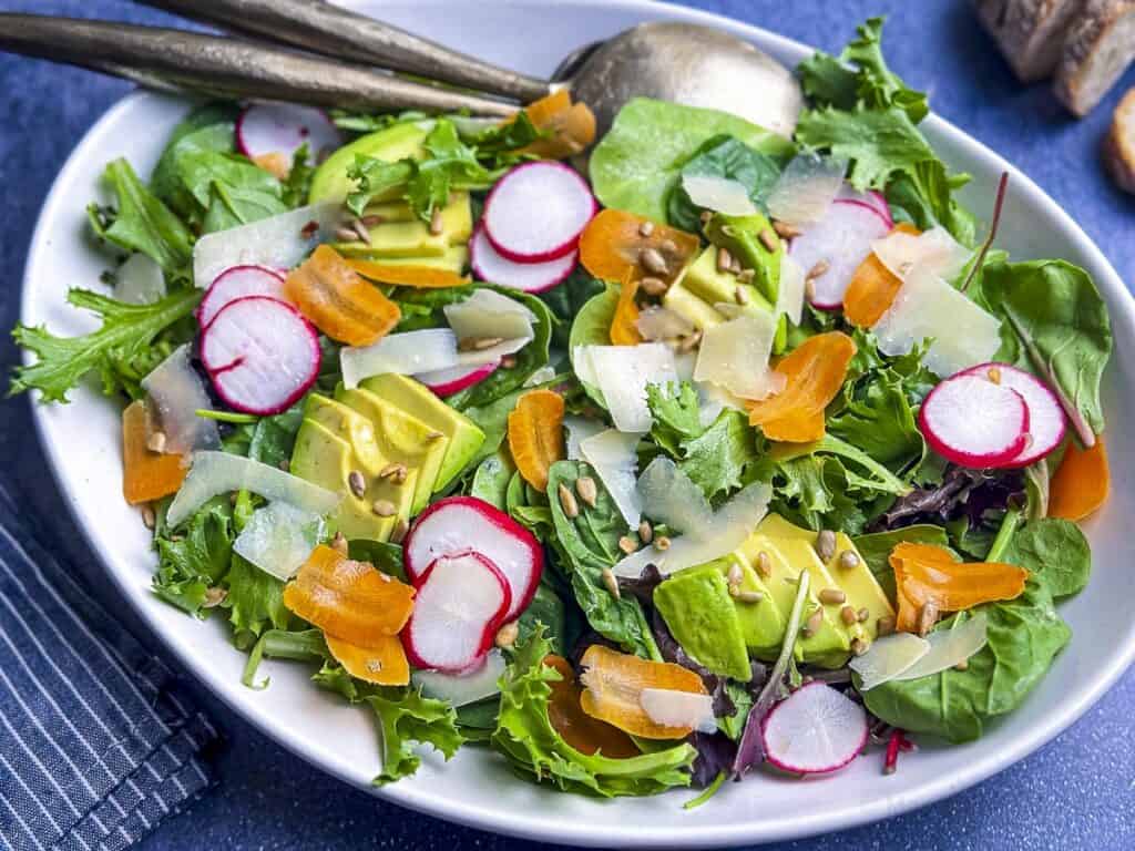 Mixed green salad with carrots, radish, and avocado on a white plate.
