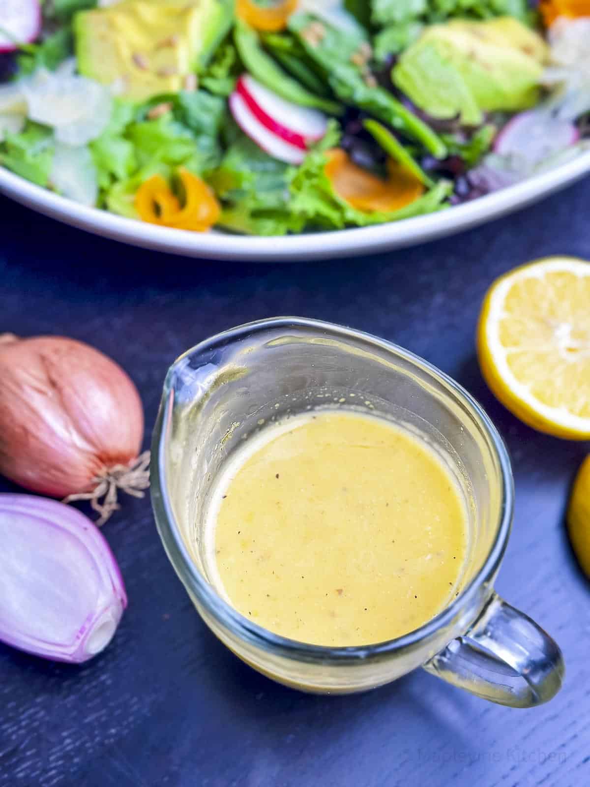 A glass pitcher filled with lemon shallot vinaigrette with lemon and shallots on the side.