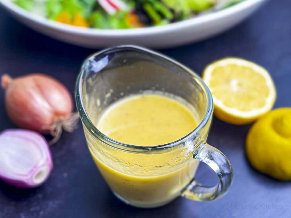 A glass pitcher filled with creamy lemon vinaigrette with lemon and shallots on the side.
