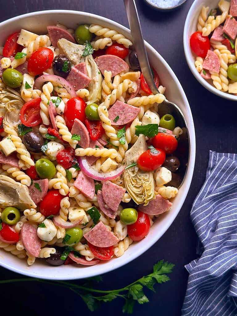Colorful antipasto pasta salad with cherry tomatoes, olives, salami, and artichokes in a white bowl.