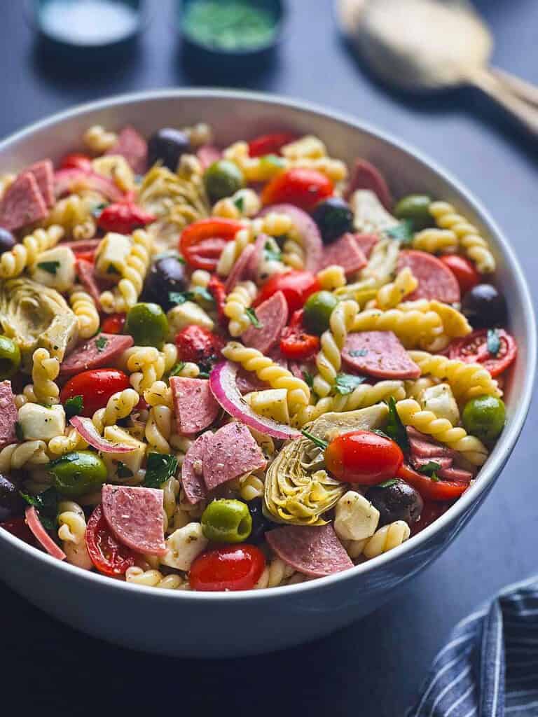 Colorful Italian pasta salad with vegetables, salami, and cheese in a white bowl.