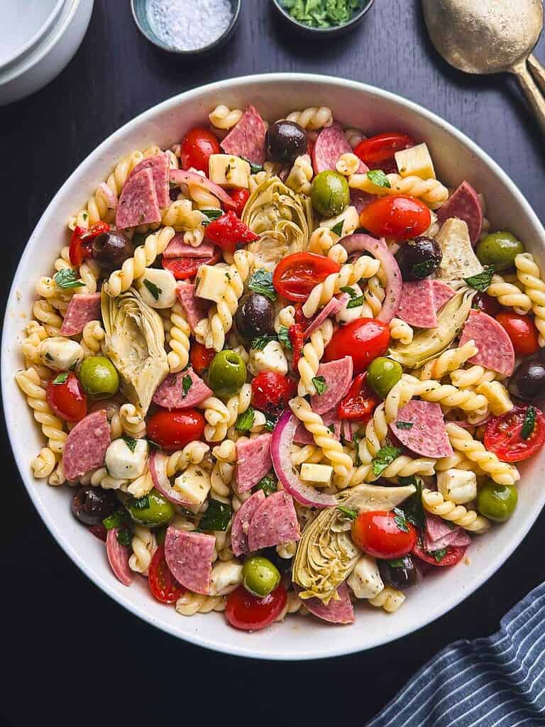 Gemelli pasta with salami, cheese, olive, tomatoes, peppers, and artichoke hearts in a white serving bowl.