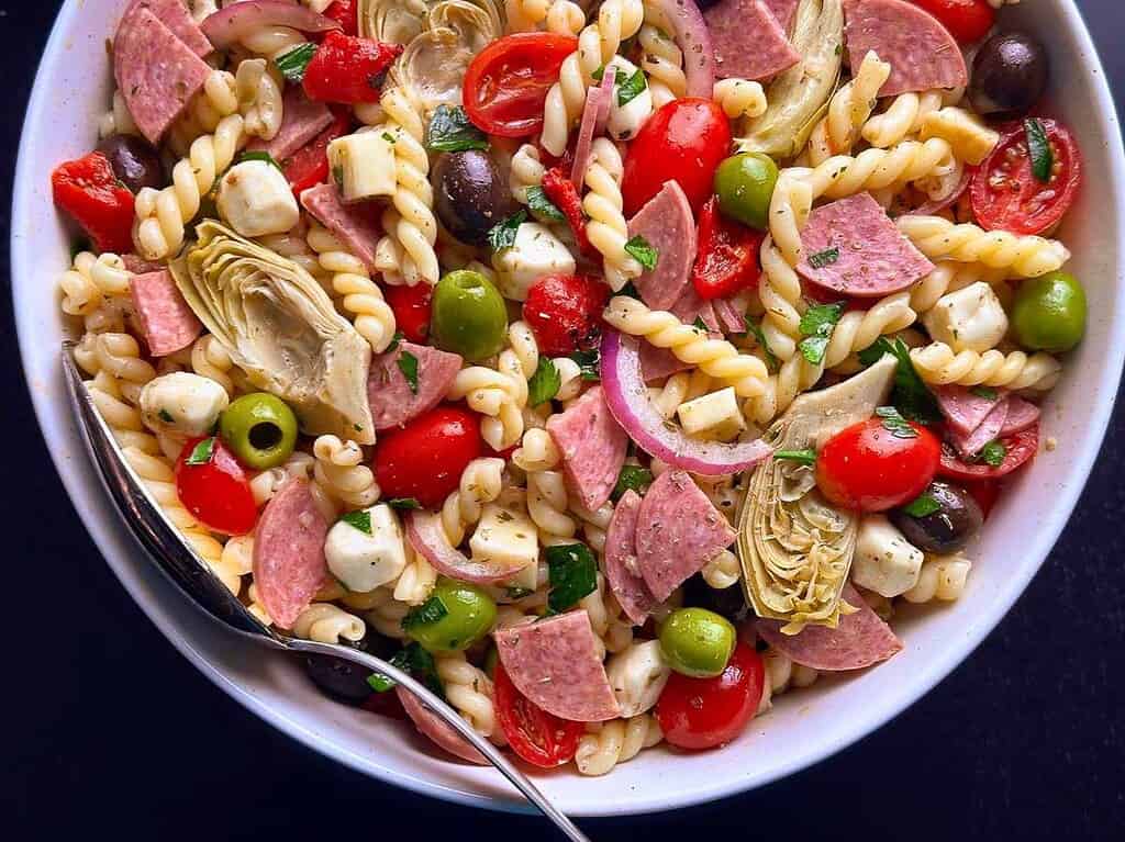 A vibrant pasta salad featuring spiral pasta, cherry tomatoes, olives, artichokes, and salami slices, ideal for picnics and casual dining.
