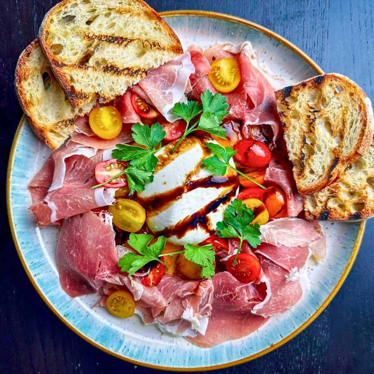 Burrata and prosciutto on a stylish plate with heirloom cherry tomatoes and fresh parsley with a balsamic glaze drizzle.