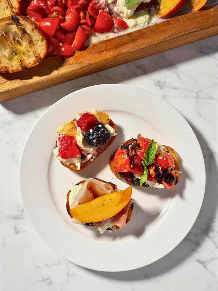 Summer crostini featuring burrata cheese, bruschetta tomatoes, peaches, prosciutto, berries, basil, and grilled crostini.