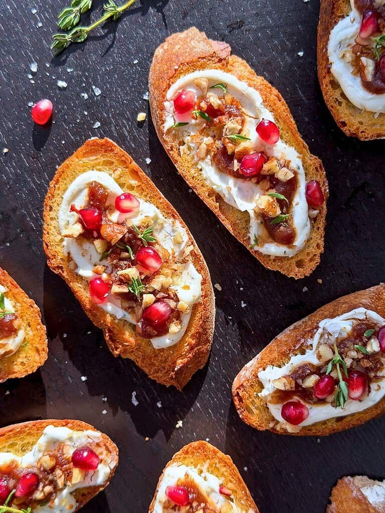 Gourmet crostini with goat cheese, apple butter, pomegranate arils, and fresh thyme on a dark background.