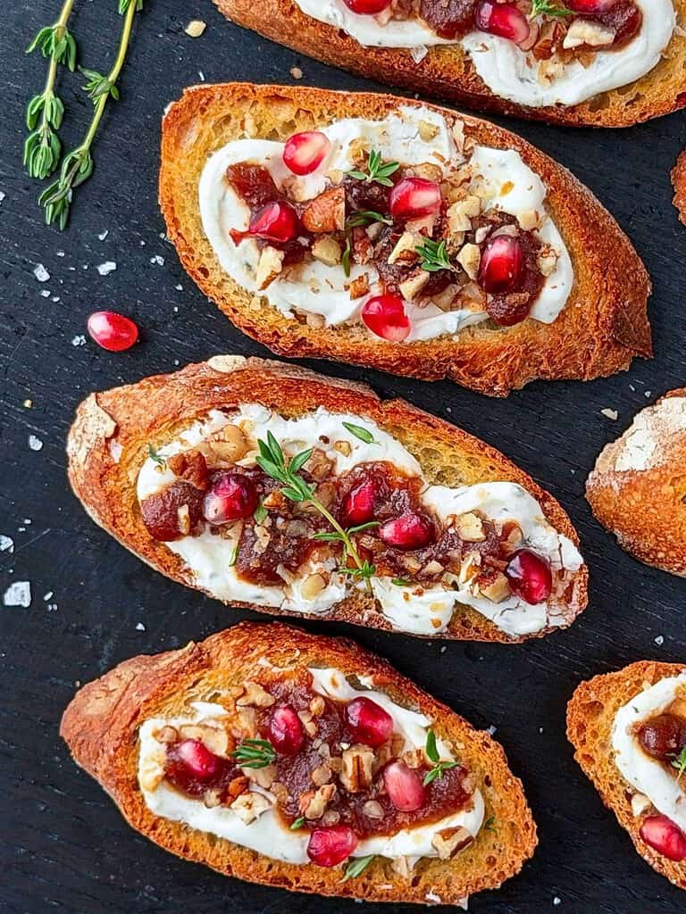 Goat cheese crostini with pomegranate, pecans, apple butter, and fresh thyme on a dark tabletop.