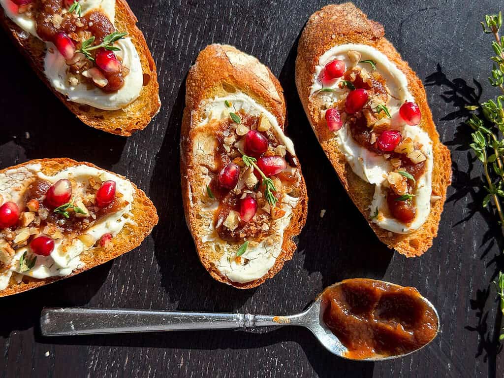 Goat cheese crostini with pomegranate, pecans, apple butter, and fresh thyme with a spoon containing apple butter.