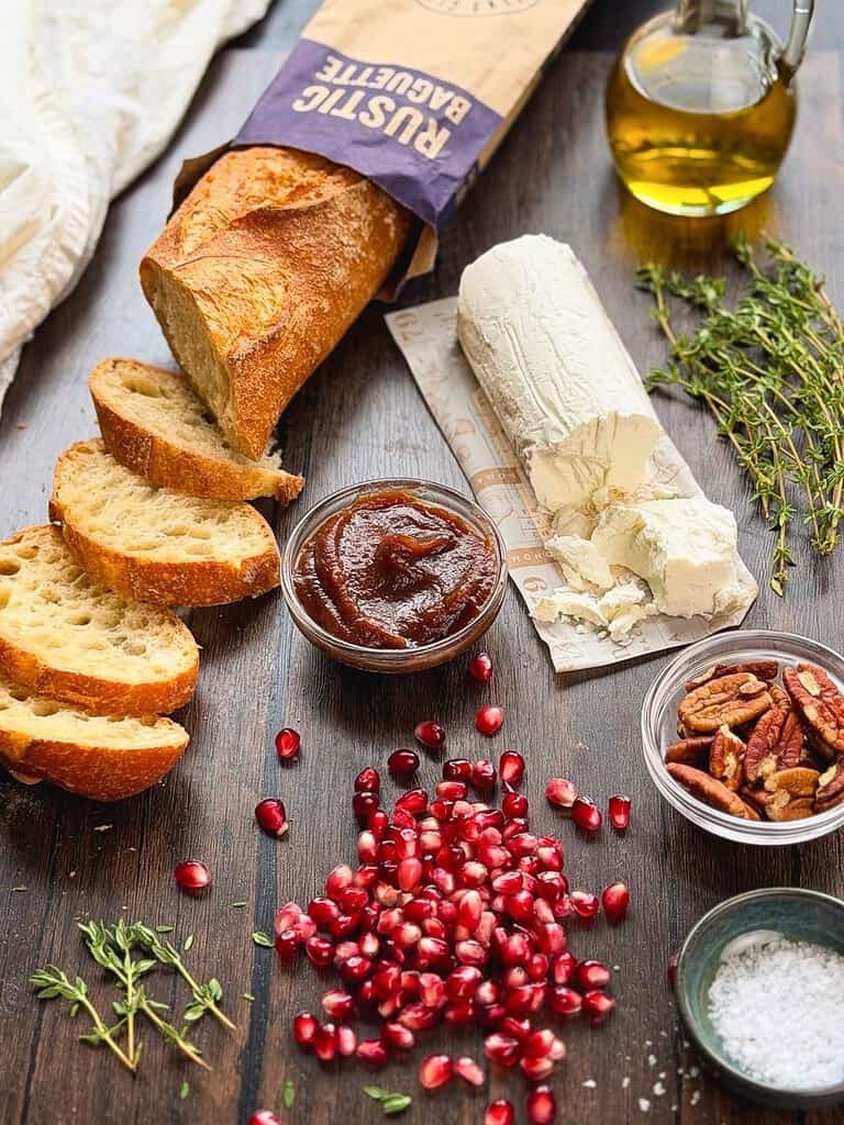 Fresh baguette, goat cheese, apple butter, pecans, pomegranate arils, and fresh thyme on a rustic table.
