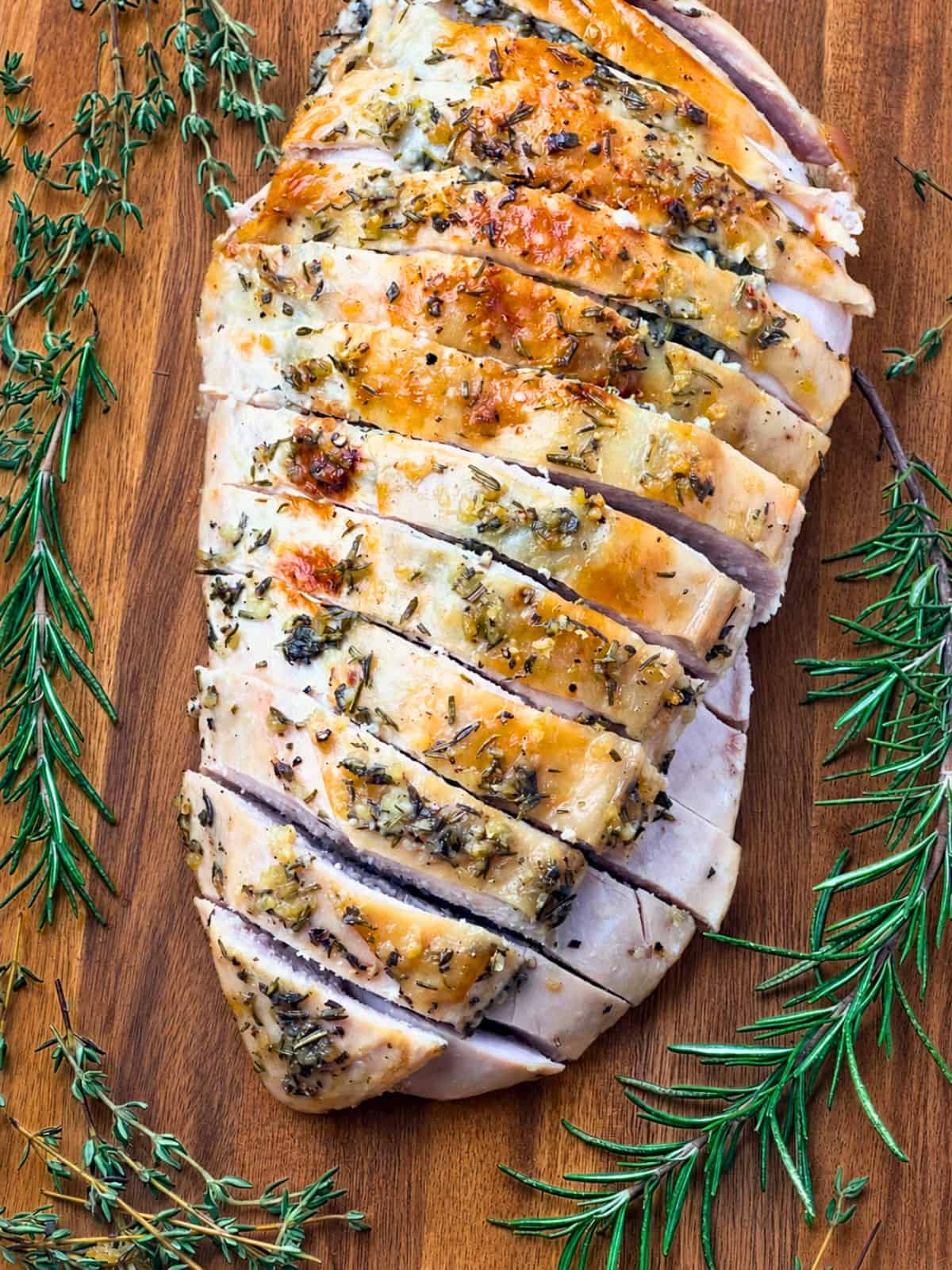 A whole roasted turkey breast sliced on a wooden board and surrounded by fresh rosemary and thyme sprigs.