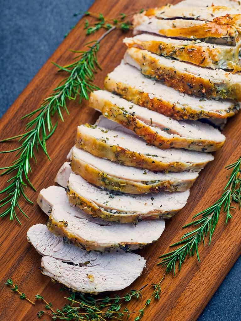 Sliced herb roasted turkey breast on a wooden board with fresh herbs.