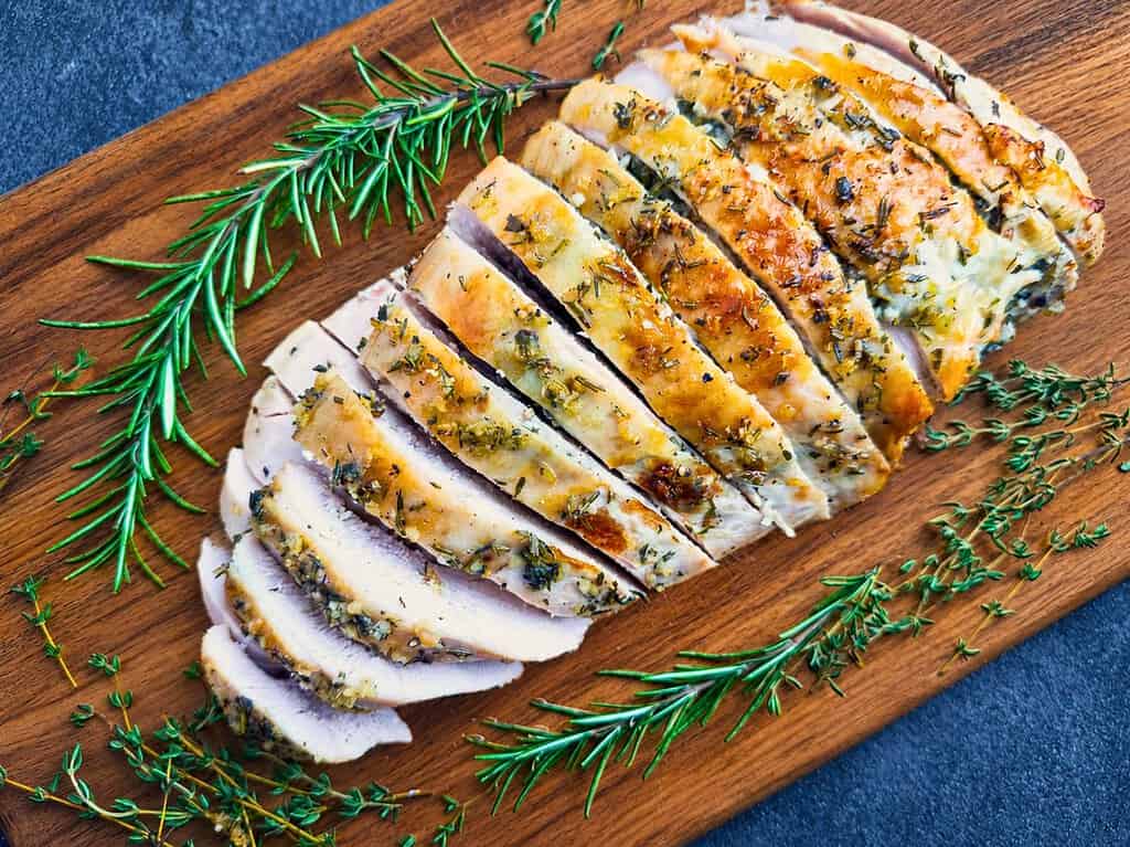 Sliced roasted turkey breast on a wooden board with fresh rosemary and thyme.
