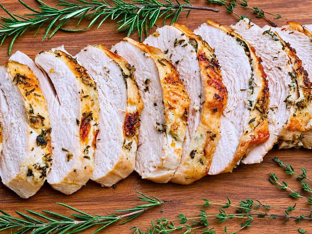 Roasted boneless turkey breast slices on a wooden board surrounded by fresh herbs.