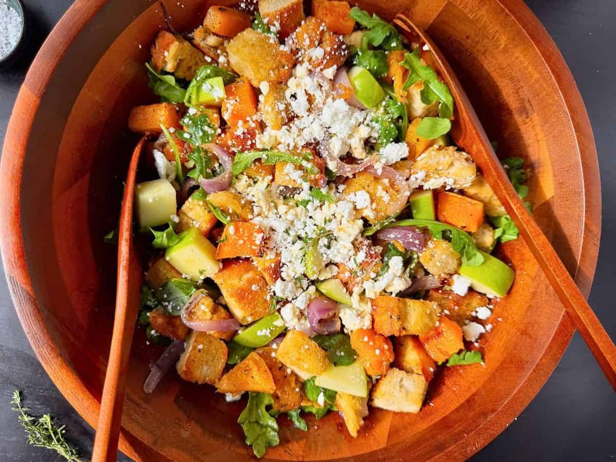 Fall panzanella salad with squash, apples, onions, arugula, and goat cheese in a large wooden bowl.