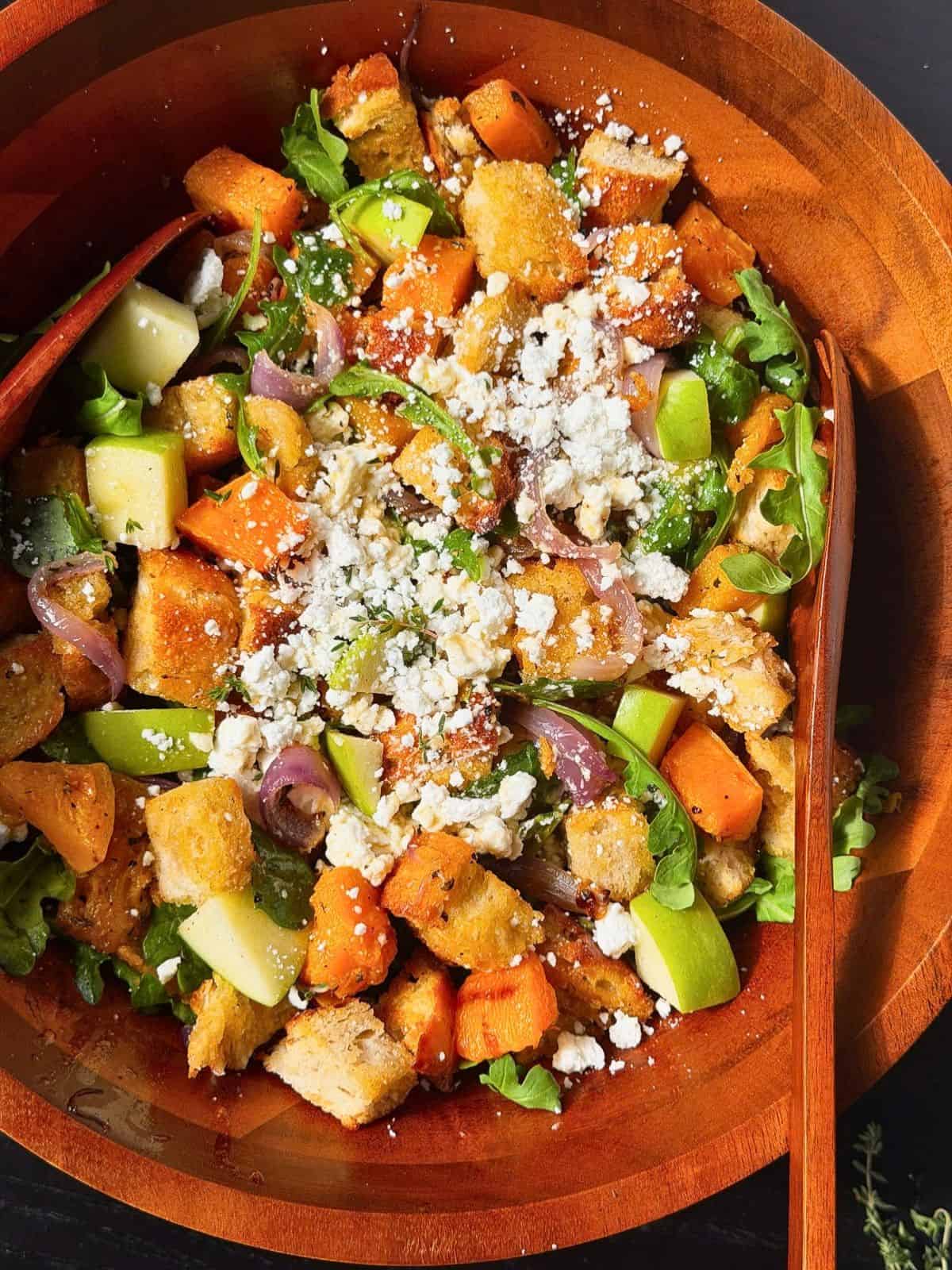 Fall panzanella salad with squash, apples, onions, arugula, and goat cheese in a large wooden bowl.