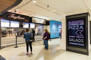 People ordering food at a mall food court with digital menu boards and signage.