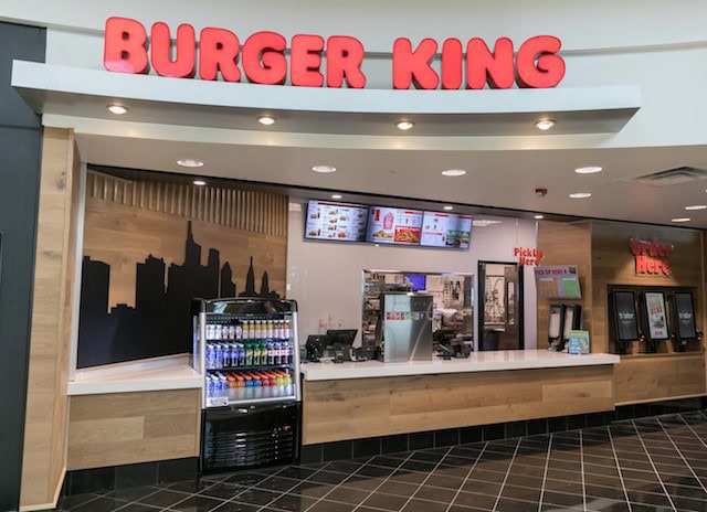 Burger King counter with menu screens and beverage fridge in mall food court.