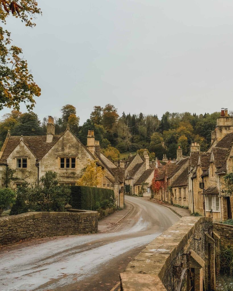 This is the idyllic English village of Castle Combe located in the Cotswolds. The village features honey-colored stone houses, thatched roofs, cascading foliage, and storybook charm. It also has a bridge over the River Bybrook as seen here in the foreground that contributes to its magical aesthetic.