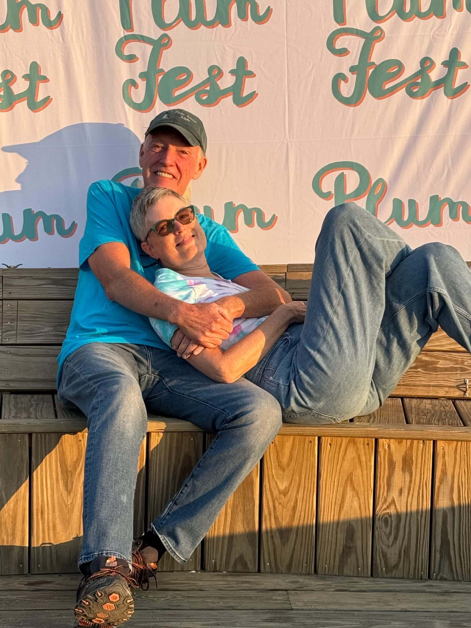 Two people sitting on wooden steps, smiling, at an event with 'Plum Fest' banner in background.