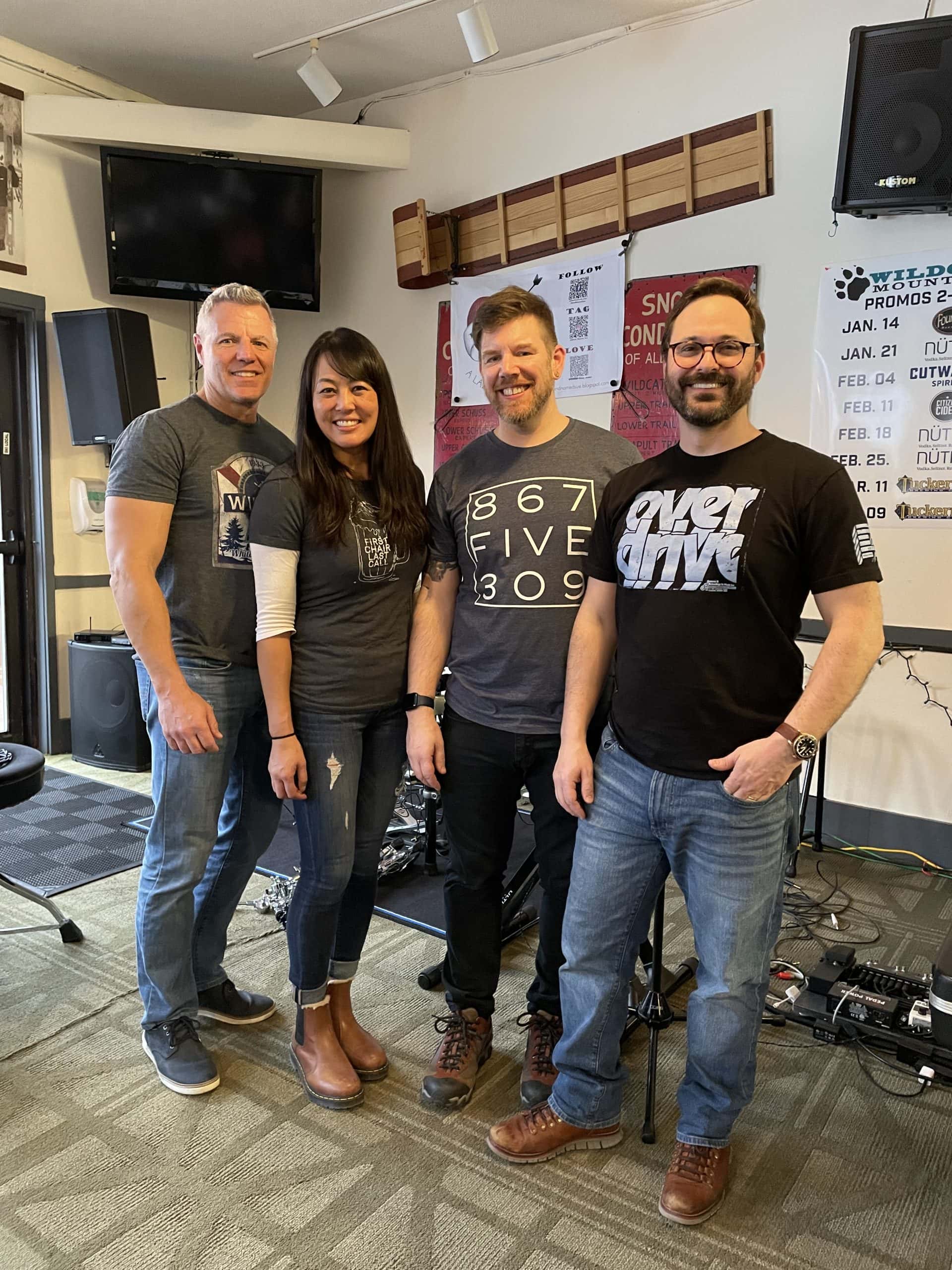 Four people standing together in a room with musical equipment and a TV.