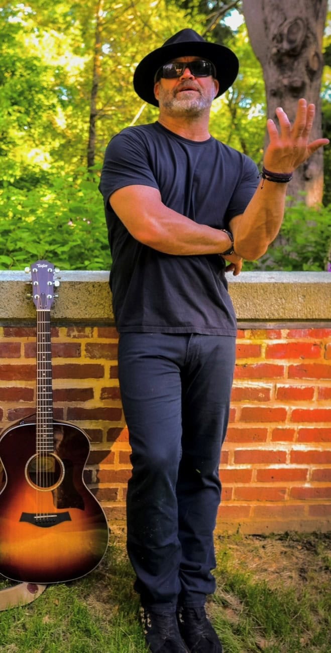 Man in black hat and sunglasses standing outdoors near a brick wall with a guitar beside him.