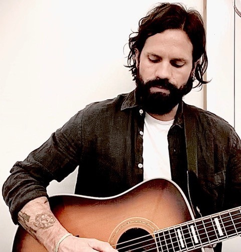 Man with dark hair and beard playing acoustic guitar indoors.