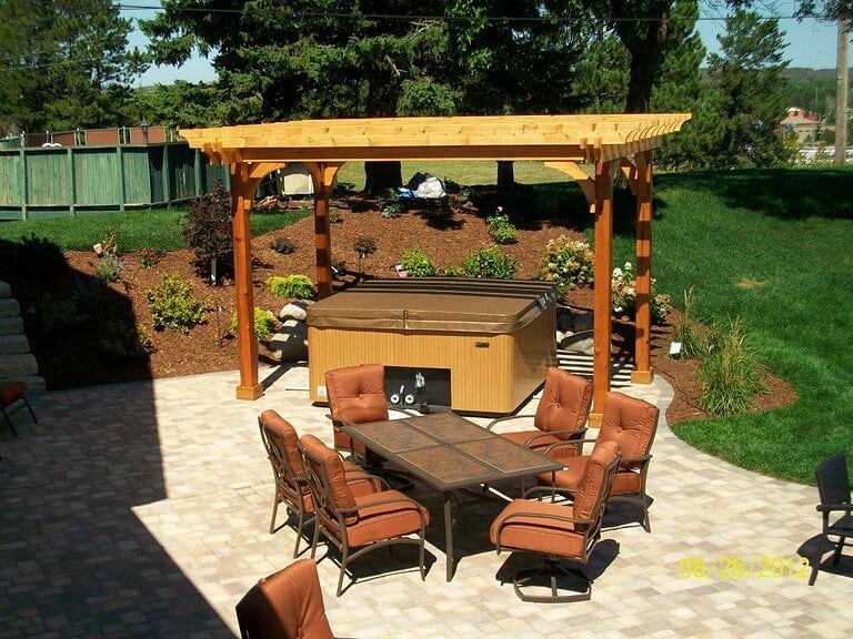 Outdoor dining area with a hot tub under a wooden pergola in a landscaped backyard.