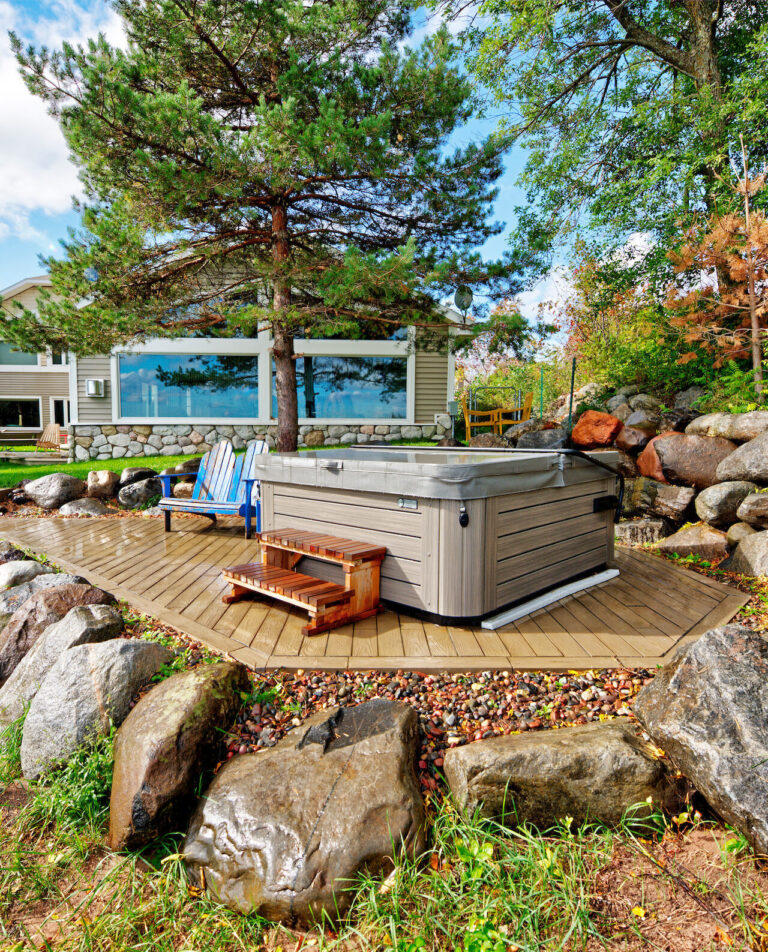 Hot tub on wooden deck surrounded by rocks and trees in backyard.
