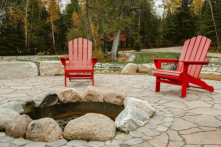 Fire pit with two red Adirondack chairs on a stone patio in a wooded backyard.