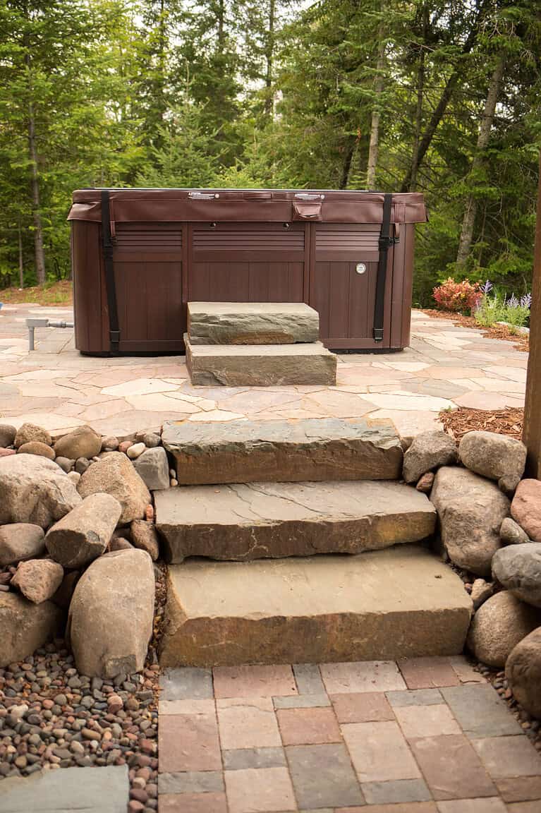 Hot tub on stone patio surrounded by trees and landscaping.