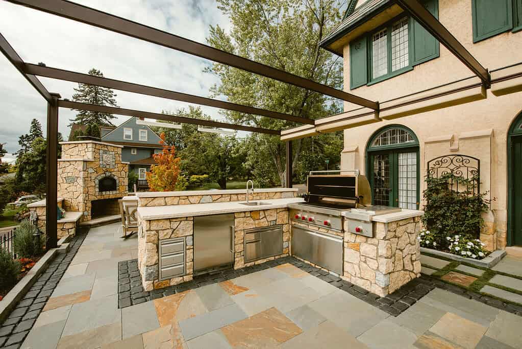 Outdoor kitchen with stone countertops, grill, and pergola in a backyard setting.