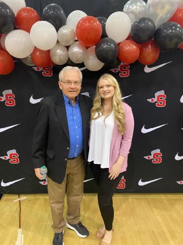 An older man and a young woman standing in a gym, smiling in front of a balloon arch in black, red, and white colors.