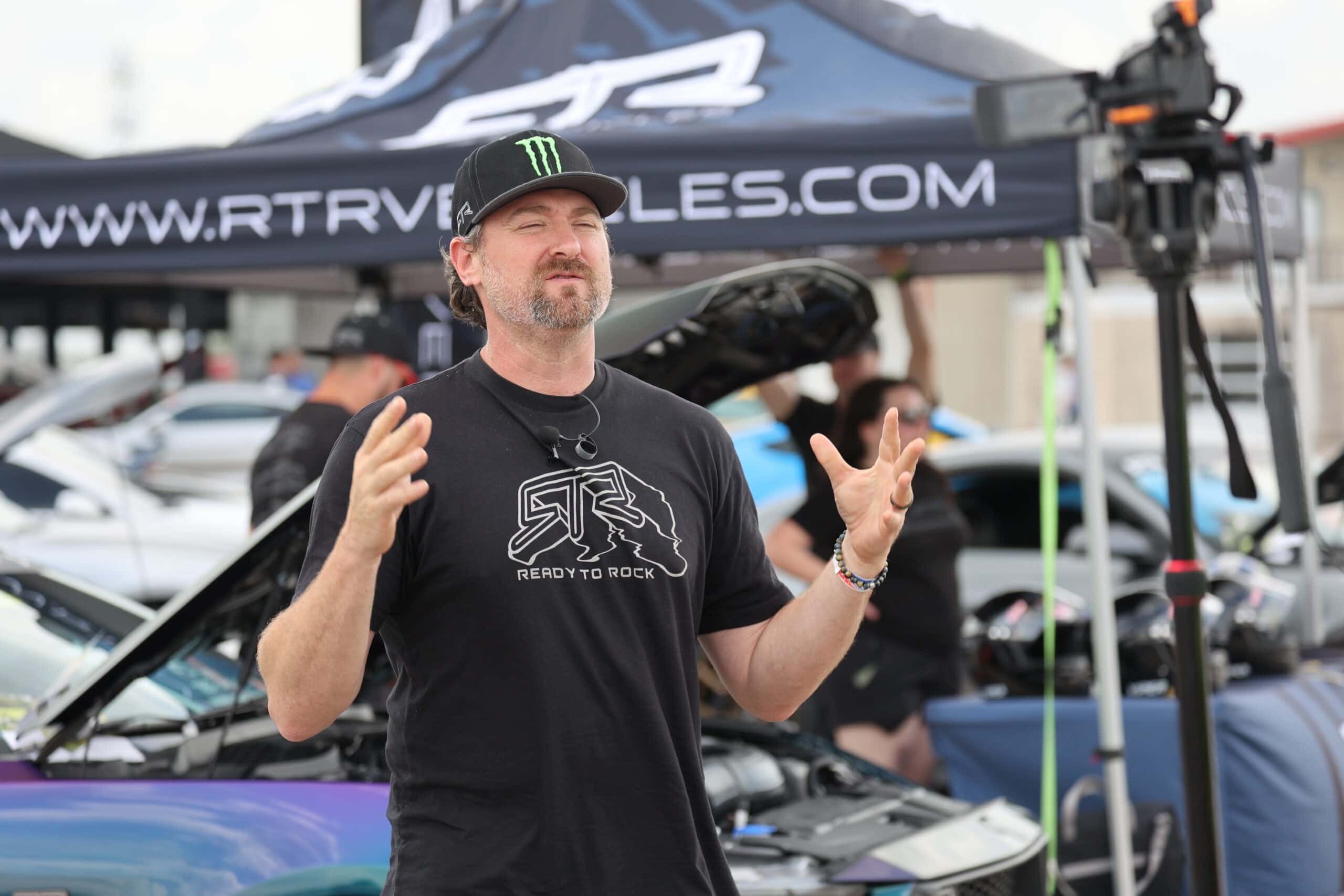Man speaking at Mustang Week car event with tents and cars in background.