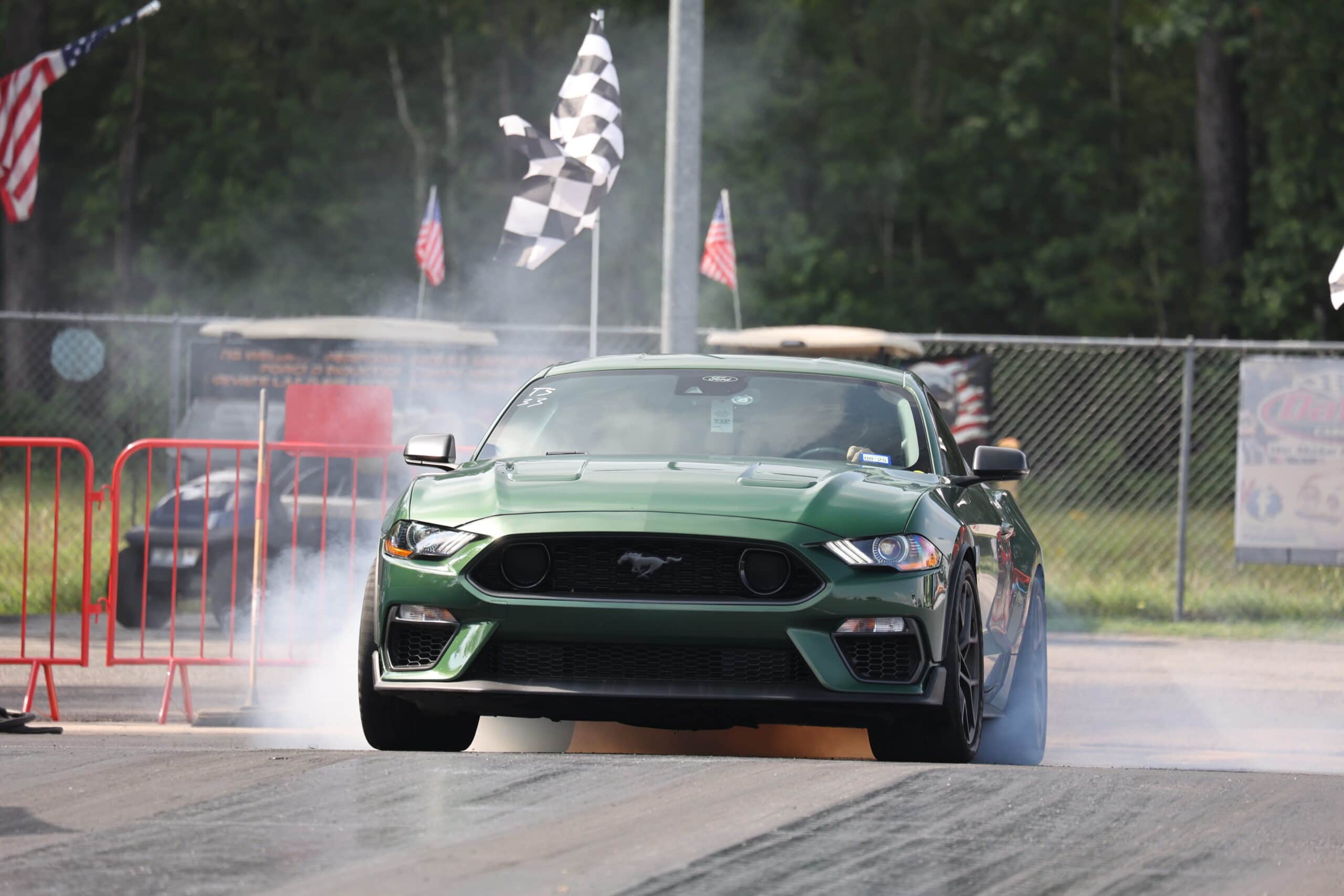 Green Ford Mustang drifting at Mustang Week event with smoke and flags.