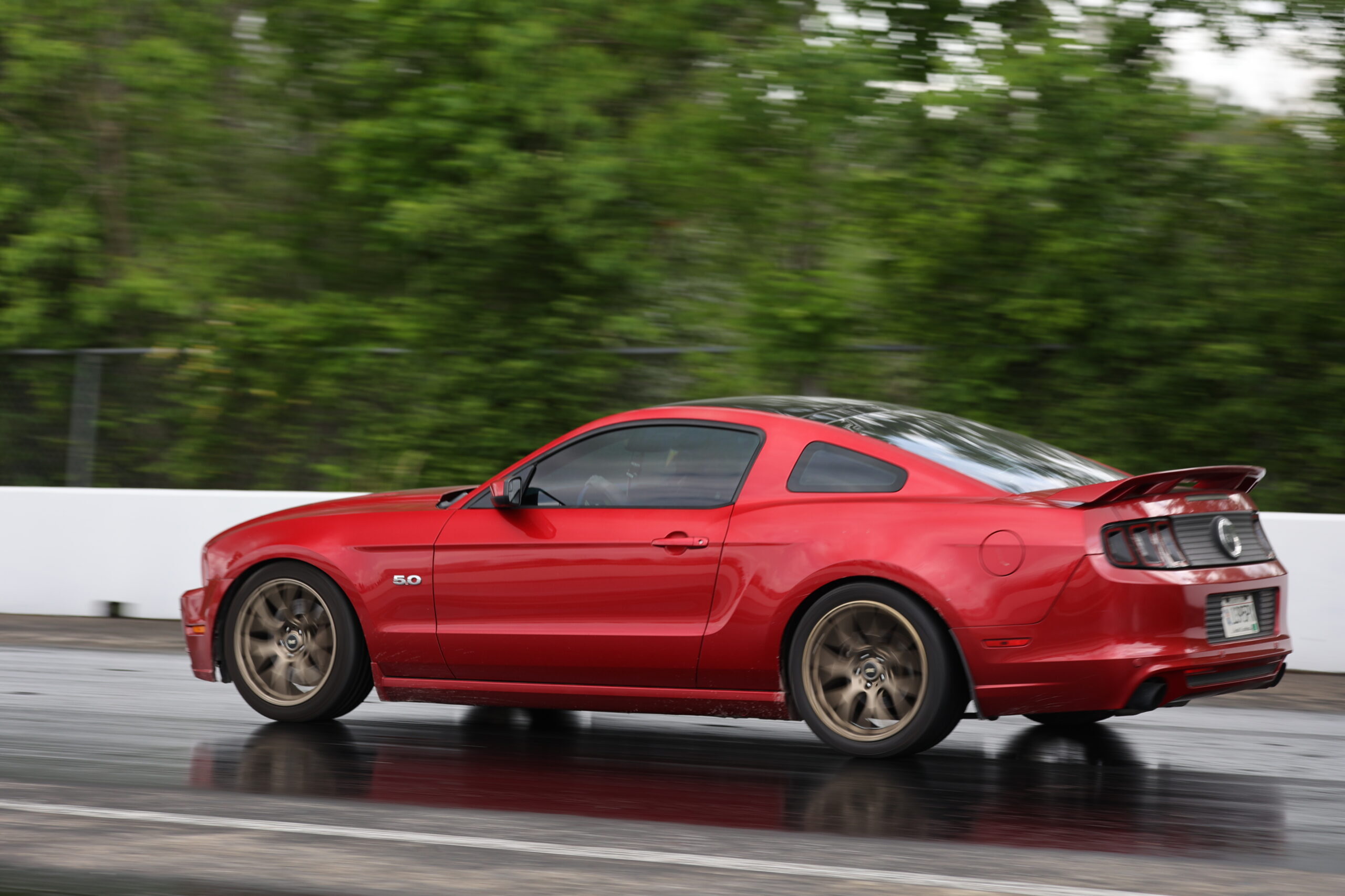 Red Ford Mustang racing on wet track during Mustang Week event.