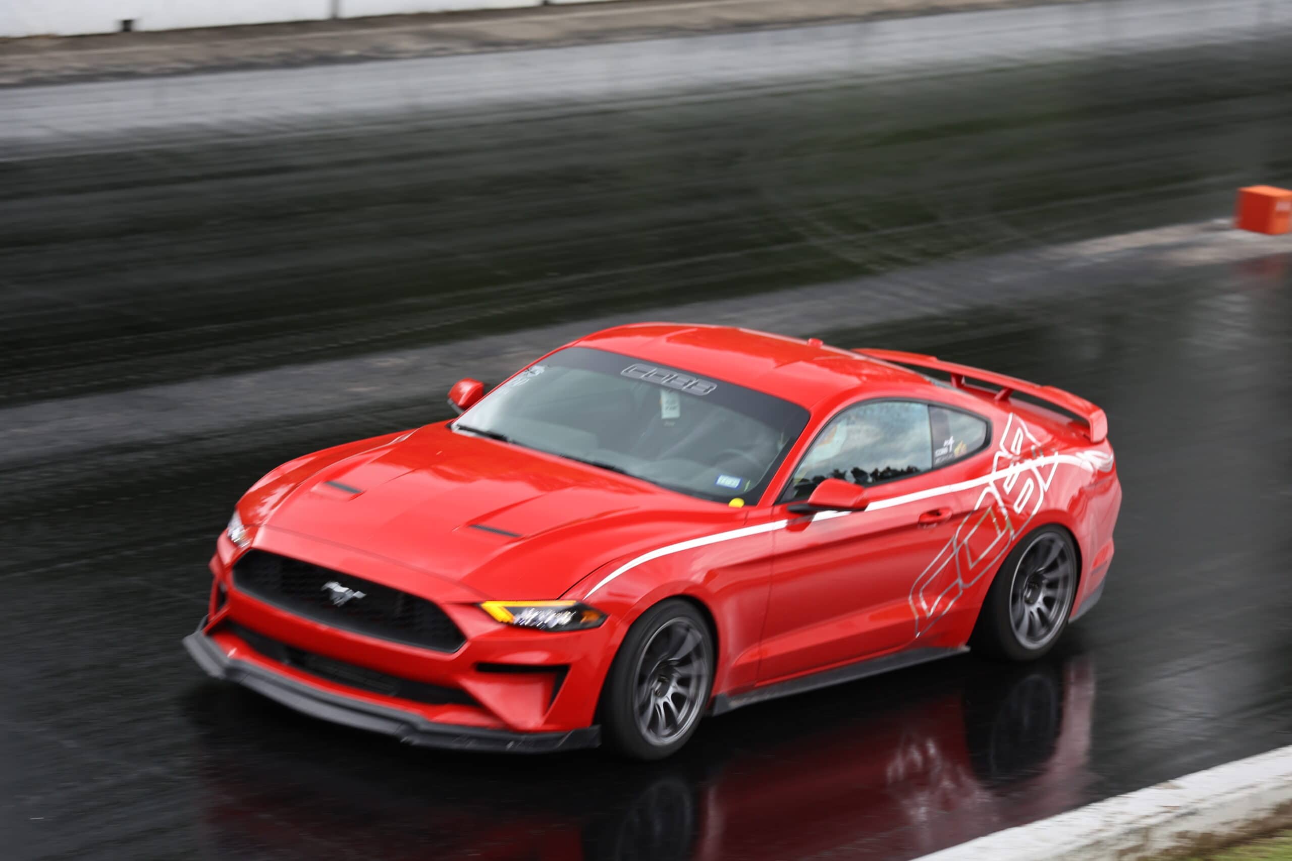 Red Ford Mustang racing on wet track at Mustang Week event.